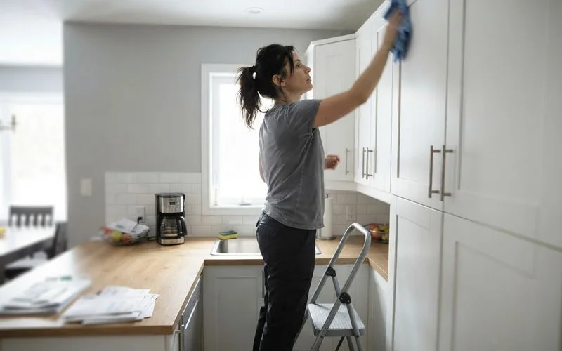 A cleaner wiping fine construction dust from the top of a kitchen cabinet after renovation work